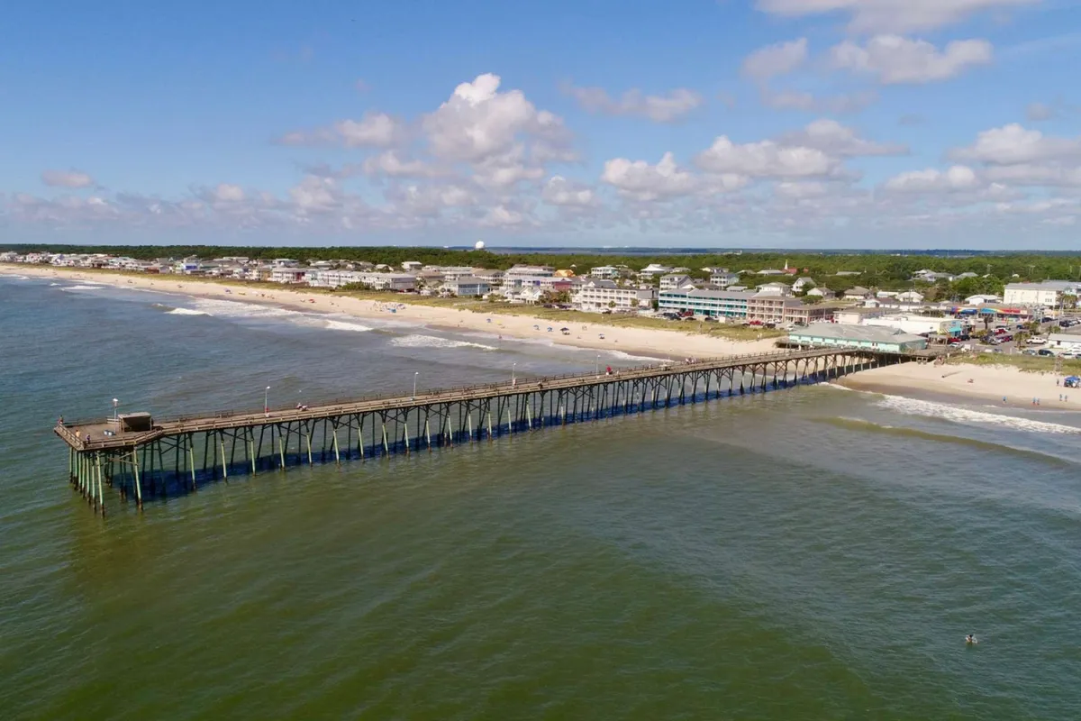 A lively exterior view featuring the iconic pier stretching into the ocean with sunlit waves, sandy beach, and cheerful visitors enjoying various beach activities. Coastal buildings sit in the distance, enhancing the sense of a vibrant yet relaxing seaside community.