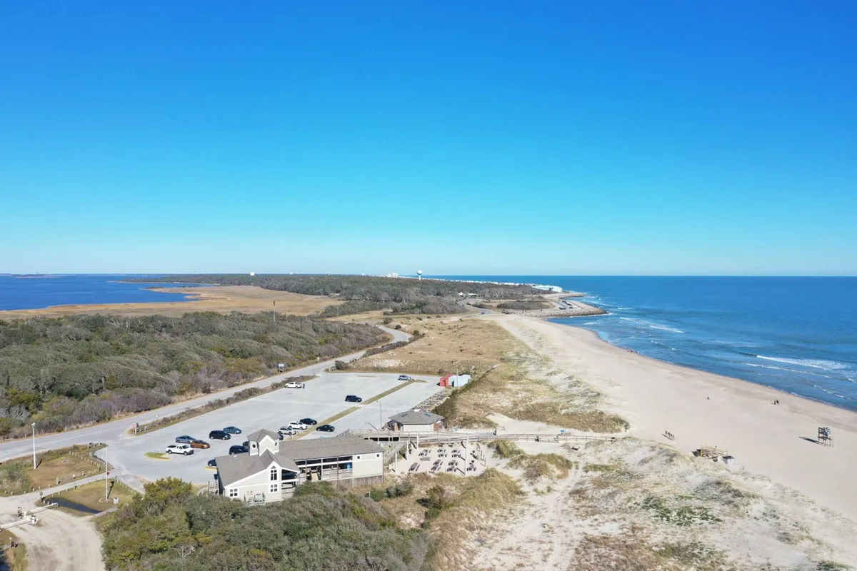 A wide-angle view of the beach with the ocean, parking area, and a nearby restaurant or gathering spot visible. The space is lively yet relaxed, showcasing the proximity of dining and entertainment options to the shore.