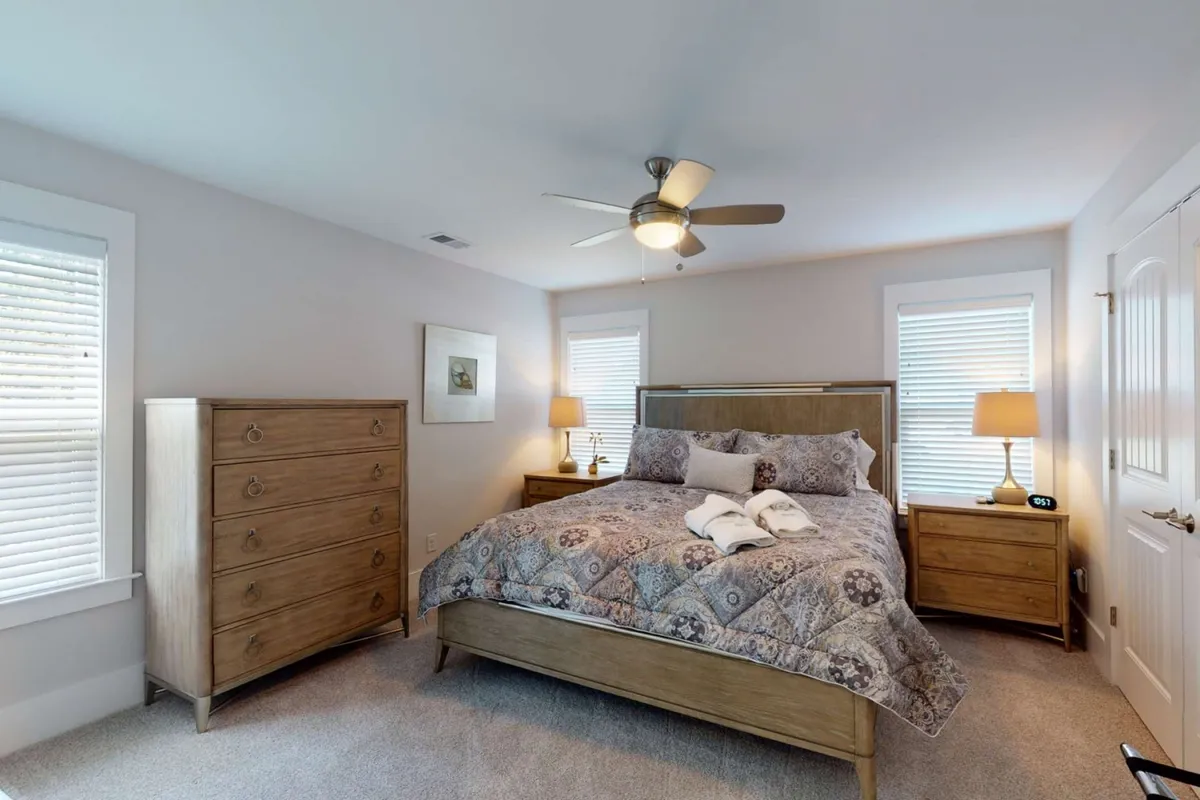 A serene queen bedroom features a bed with a patterned quilt, nightstands with lamps, a wooden dresser, and artwork on the wall. The mood is calm and inviting, ideal for couples or friends seeking comfort and privacy.