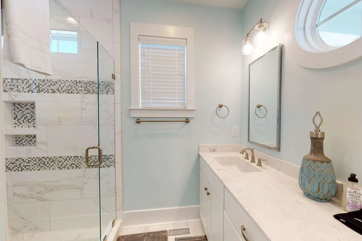 A luxurious bathroom with a glass-door shower, double vanity, a round window letting in morning light, and decorative touches like a vase. The setting is serene, spa-inspired, and perfect for relaxation and self-care routines.