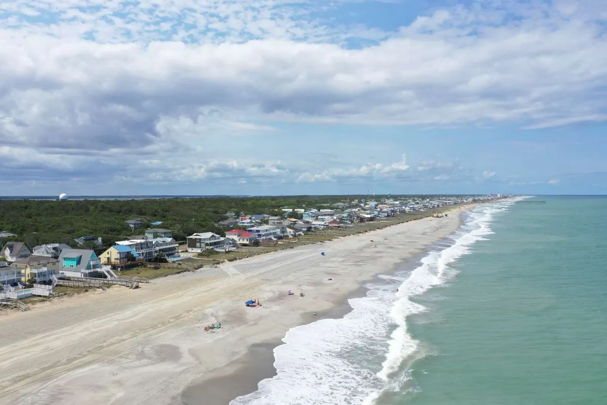 A tranquil scene of Kure Beach with gentle waves rolling onto the sandy shoreline, colorful houses in the background, and a bright, open sky above. The atmosphere exudes relaxation and the quintessential beach experience, inviting guests to enjoy sunbathing, swimming, or a leisurely stroll.