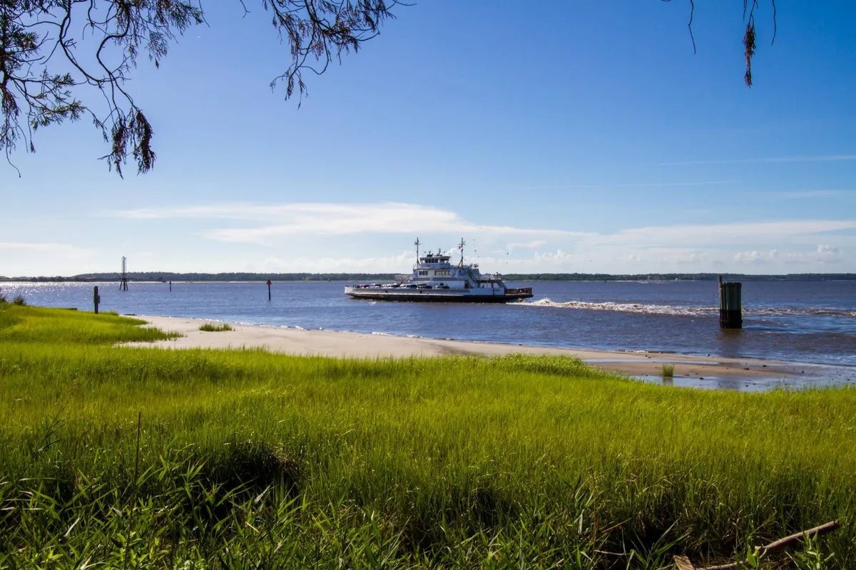 A serene view of the Fort Fisher ferry gliding across calm waters under a blue sky, with lush green grass lining the shore. This image showcases the ferry ride to Southport, a notable family adventure mentioned in the section, and hints at the broader exploration opportunities nearby.