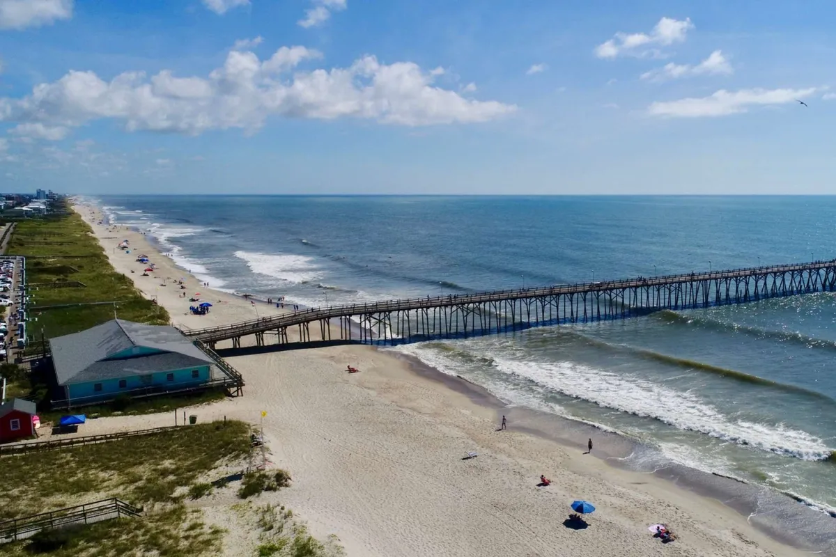 A wide shot of the Kure Beach pier stretching over the ocean, bordered by golden sand and a relaxed shoreline. The pier is bustling with people enjoying fishing, strolling, or simply taking in the view. Gentle waves roll in under a sky with scattered clouds, evoking a sense of classic beach fun and local tradition.