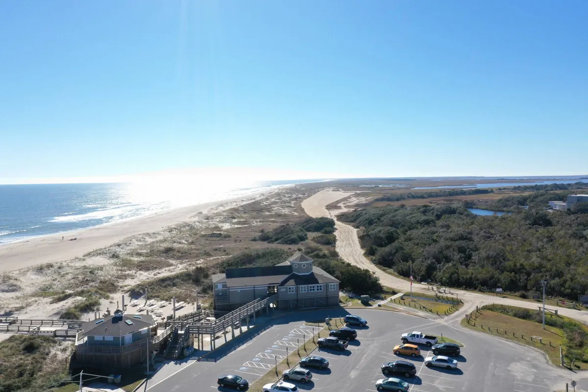 A spacious view of the beach and ocean is visible beyond a parking area and a level boardwalk, surrounded by grassy dunes. The flat terrain and clear path make accessing the shoreline inviting and manageable for all guests, including those with mobility considerations.