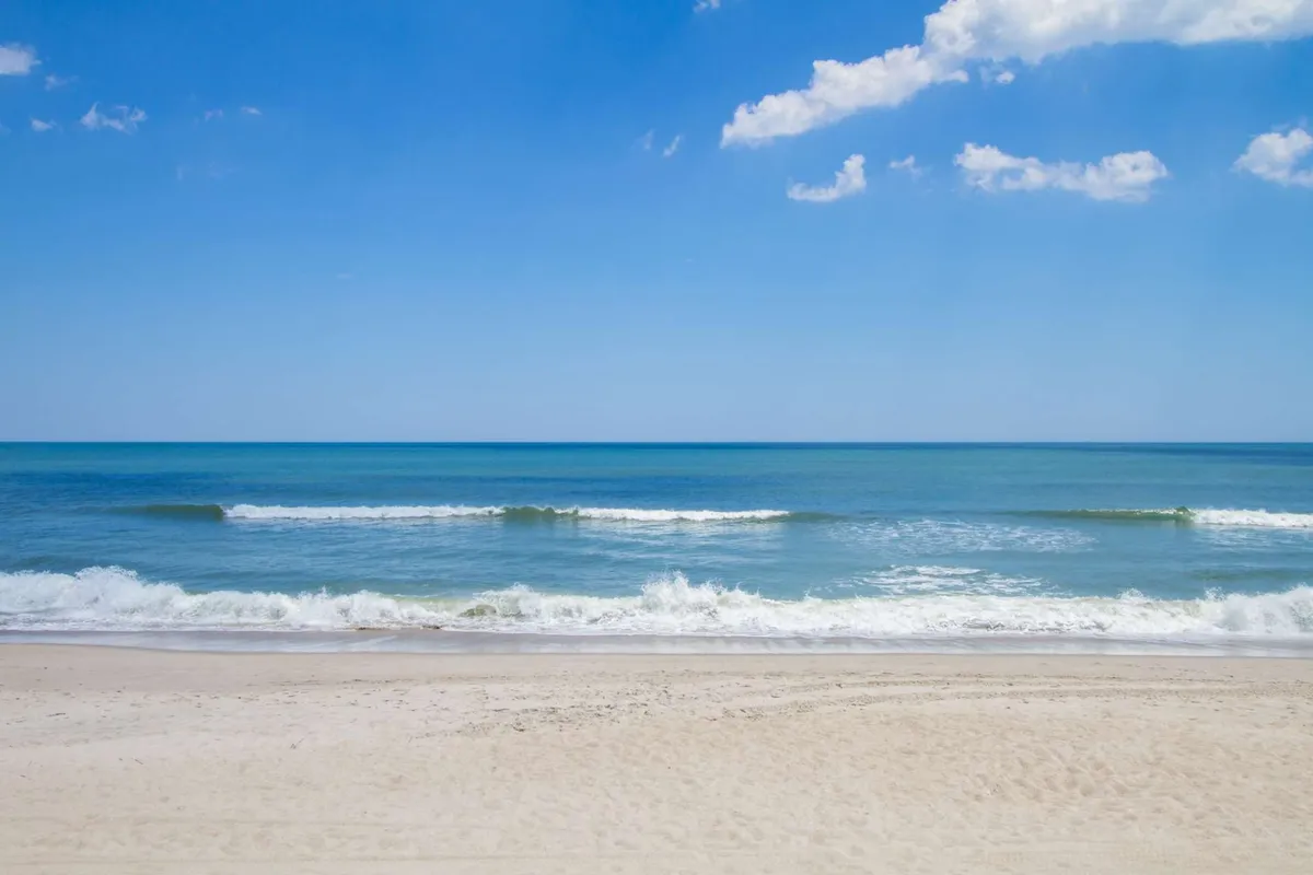 A wide, flat beach opens out to the ocean under clear skies, creating a peaceful, inviting setting. The gentle terrain and openness suggest accessibility for beach walks, sunbathing, and leisure activities for guests of all ages and mobility levels.