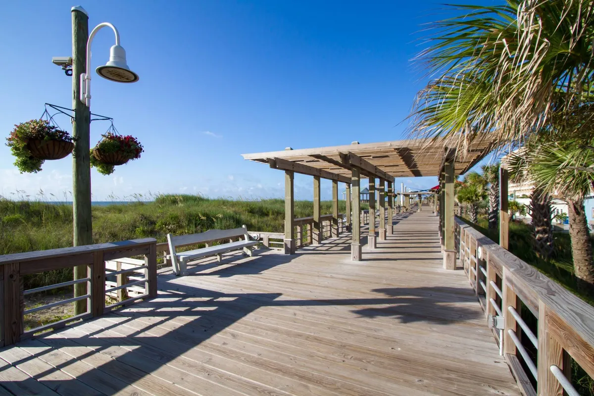 A scenic exterior view featuring a classic wooden boardwalk lined with hanging flower baskets and palm trees, leading toward the water. The canopy structure and lush surroundings evoke a welcoming coastal vibe, ideal for gatherings and strolls.