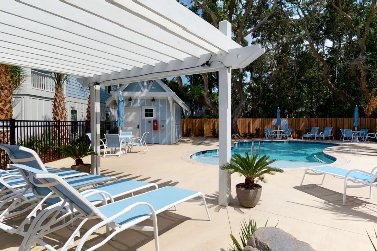 The image features an inviting, sun-drenched swimming pool surrounded by comfortable lounge chairs and a stylish pergola, set against a backdrop of lush trees in a peaceful neighborhood. The space is designed for relaxation, sunbathing, socializing, and enjoying the outdoors, with the promise of sunset swims and poolside gatherings.