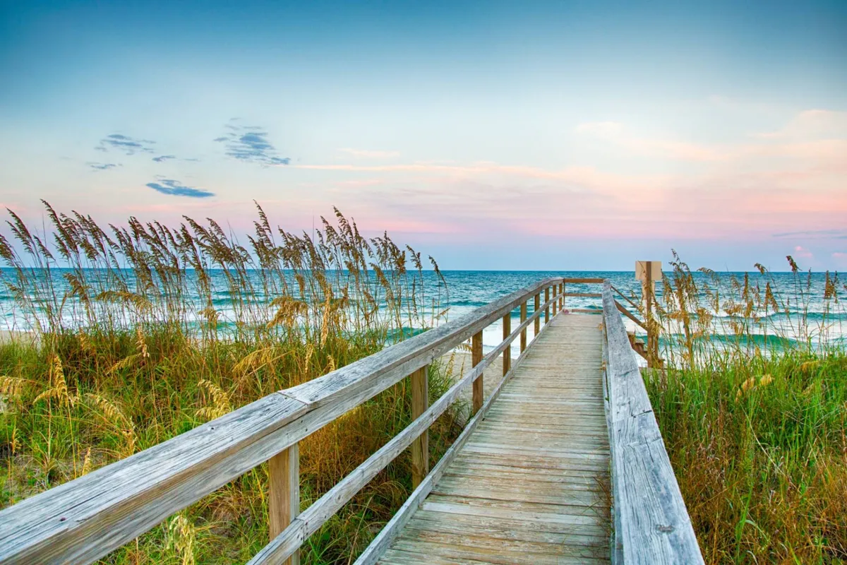 A serene view from a wooden boardwalk leading through grassy dunes towards the ocean, with gentle waves and a soft sunset sky. The natural landscape blends tranquility with anticipation, perfect for evening strolls or quiet reflection by the shore.