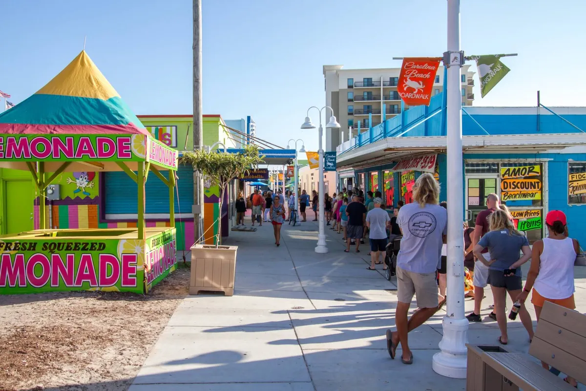 A cheerful, bustling scene at a vibrant local lemonade stand surrounded by colorful storefronts and crowds of people. Families are seen walking, shopping, and enjoying refreshments, capturing the festive, lively energy of Carolina Beach's boardwalk area.